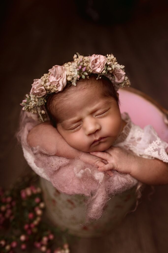 Newborn baby girl in a buck wearing a light pink floral crown, she is sleeping peacefully