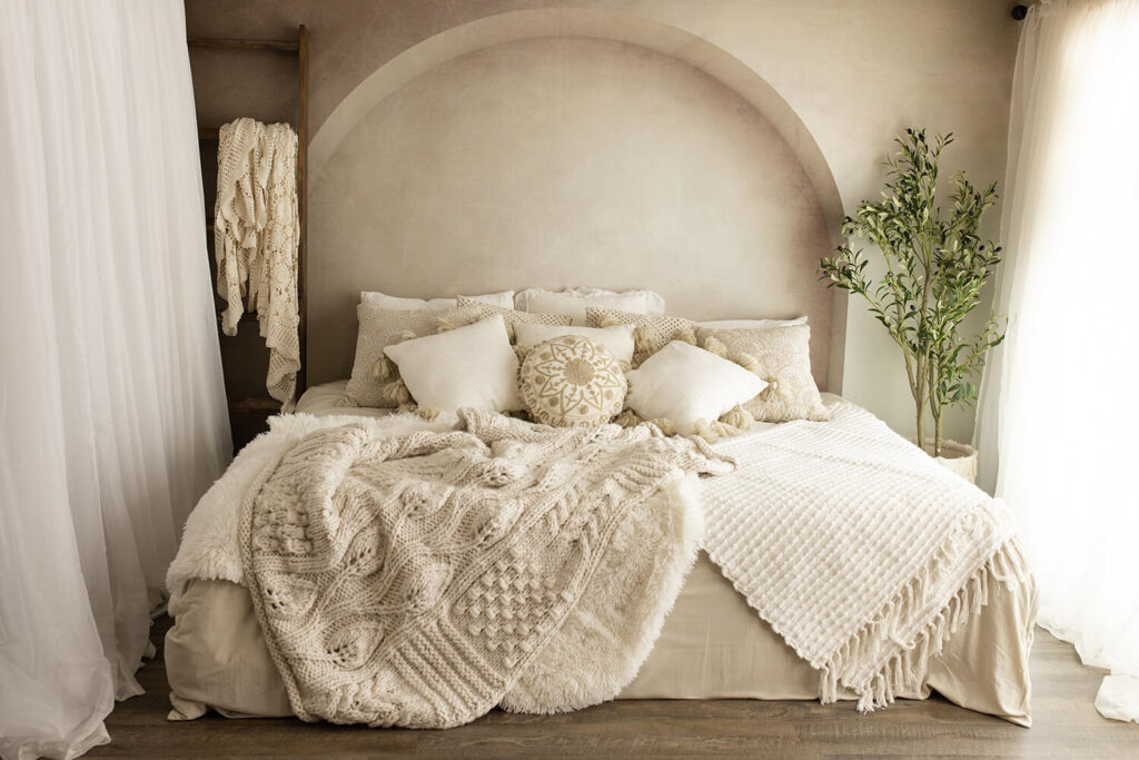 Beautiful Cream and ivory bed with lots of pillows and throw blankets in front of an arch wall with an olive tree to the right