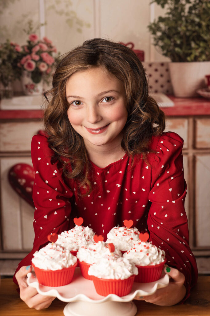 "Smiling young girl in a red dress with white heart patterns, holding a plate of festive Valentine's Day cupcakes topped with whipped cream and heart-shaped decorations. The cozy backdrop features a Valentine's-themed kitchen with warm tones and floral accents."